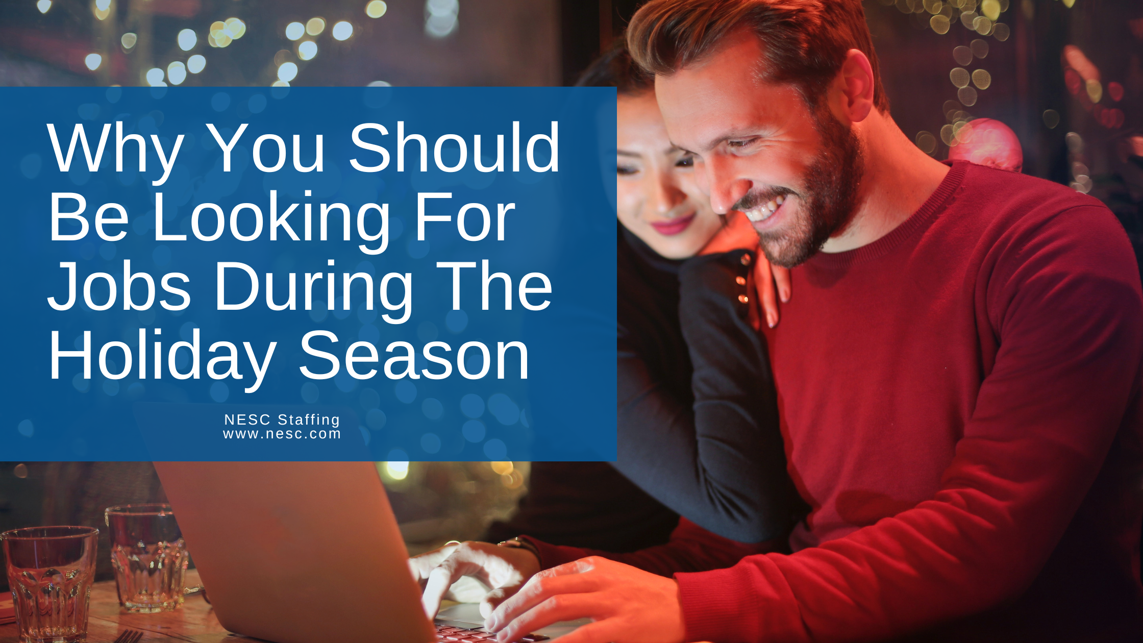 Woman watches and leans on the man, while he types on a laptop. Both of them are smiling while he applies to jobs online. Behind them are Christmas tree lights.